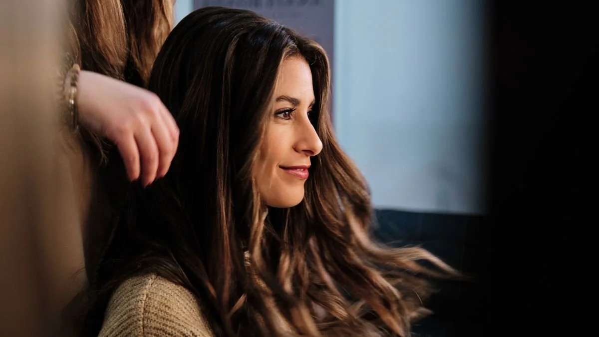 A woman with long brown hair is sitting, smiling softly, with a person behind her touching her hair. The woman is wearing a beige sweater and looking to the side.