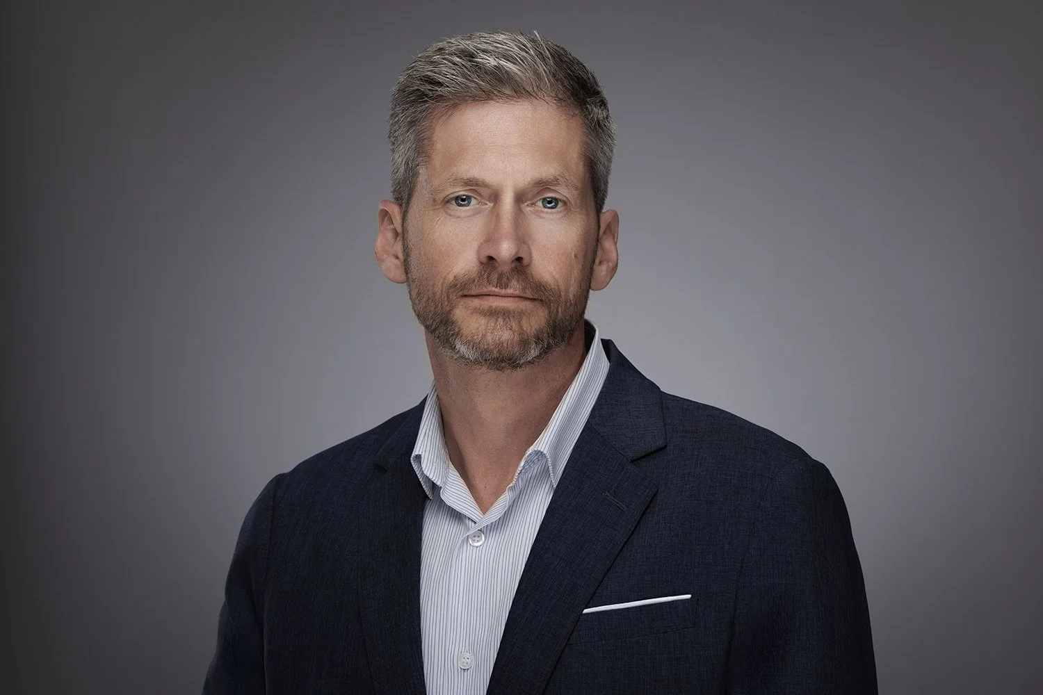 A middle-aged man with short, light brown hair and a beard, wearing a dark suit jacket and a white dress shirt, looking confidently at the camera against a grey background.