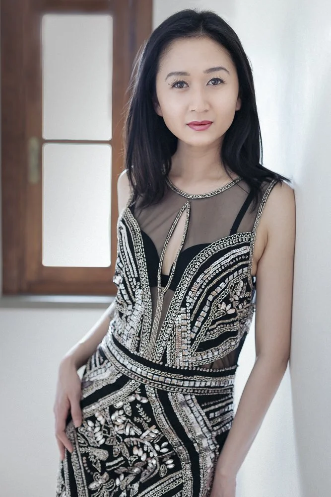 A woman with black hair wearing a black and silver beaded dress standing indoors near a window
