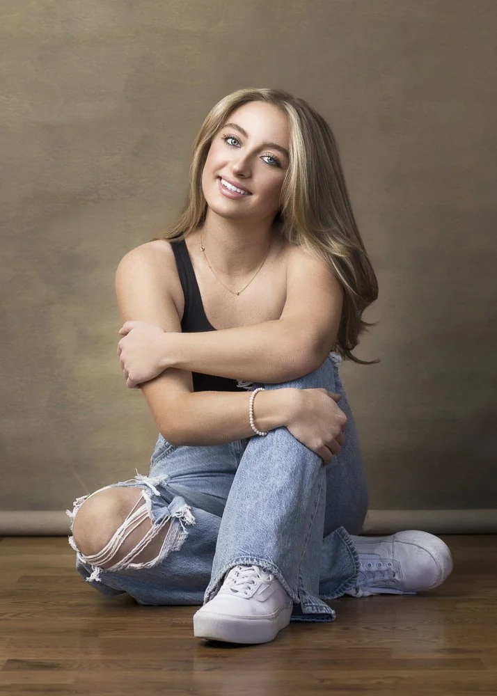 A young woman with blonde hair smiling, sitting on the floor with her arms wrapped around her knees, wearing a black tank top, ripped jeans, white sneakers, and accessorized with a pearl bracelet and a necklace, against a plain background.