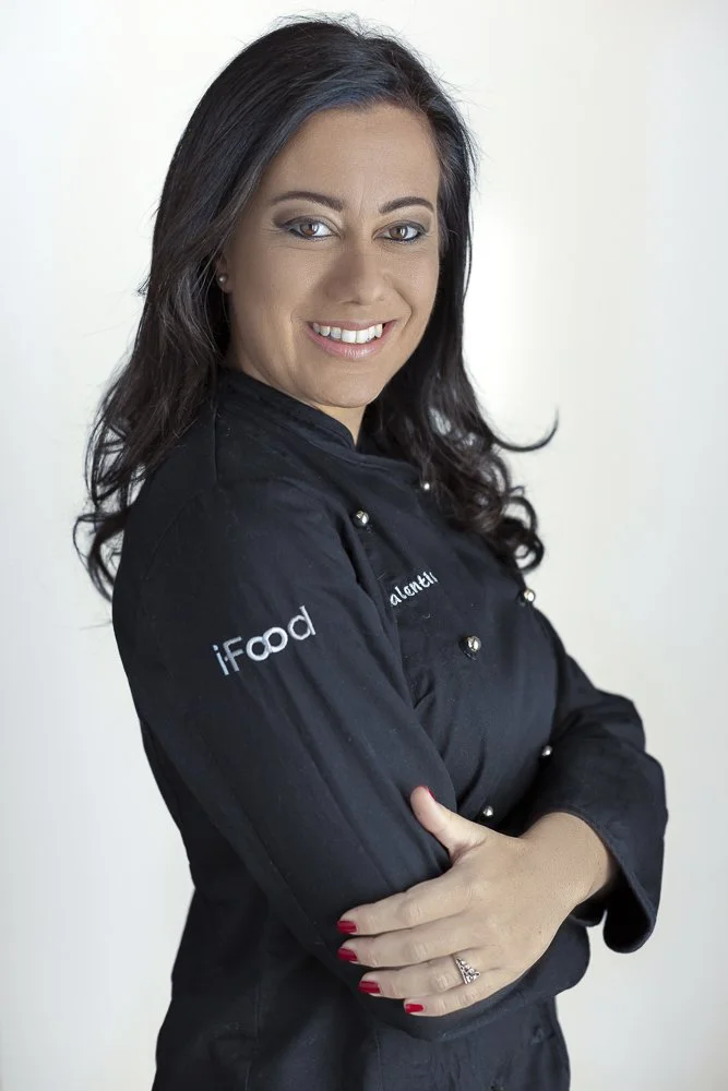 A confident woman with long dark hair and red nail polish, dressed in a black chef's jacket with 'iFood' branding, looking at the camera with a smile, arms crossed, against a plain white background.