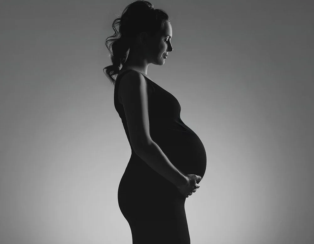 Silhouette of a pregnant woman in profile, standing with her hands on her belly, against a plain background.