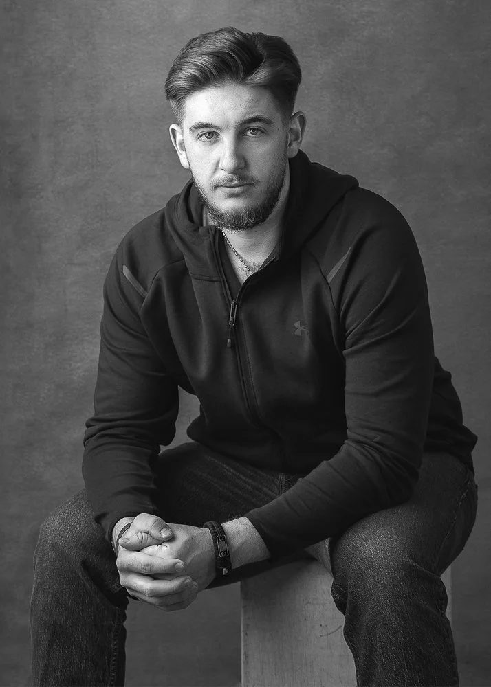A young man with light hair and a beard, wearing a dark zip-up jacket, sitting on a wooden stool against a plain backdrop in a black-and-white portrait.