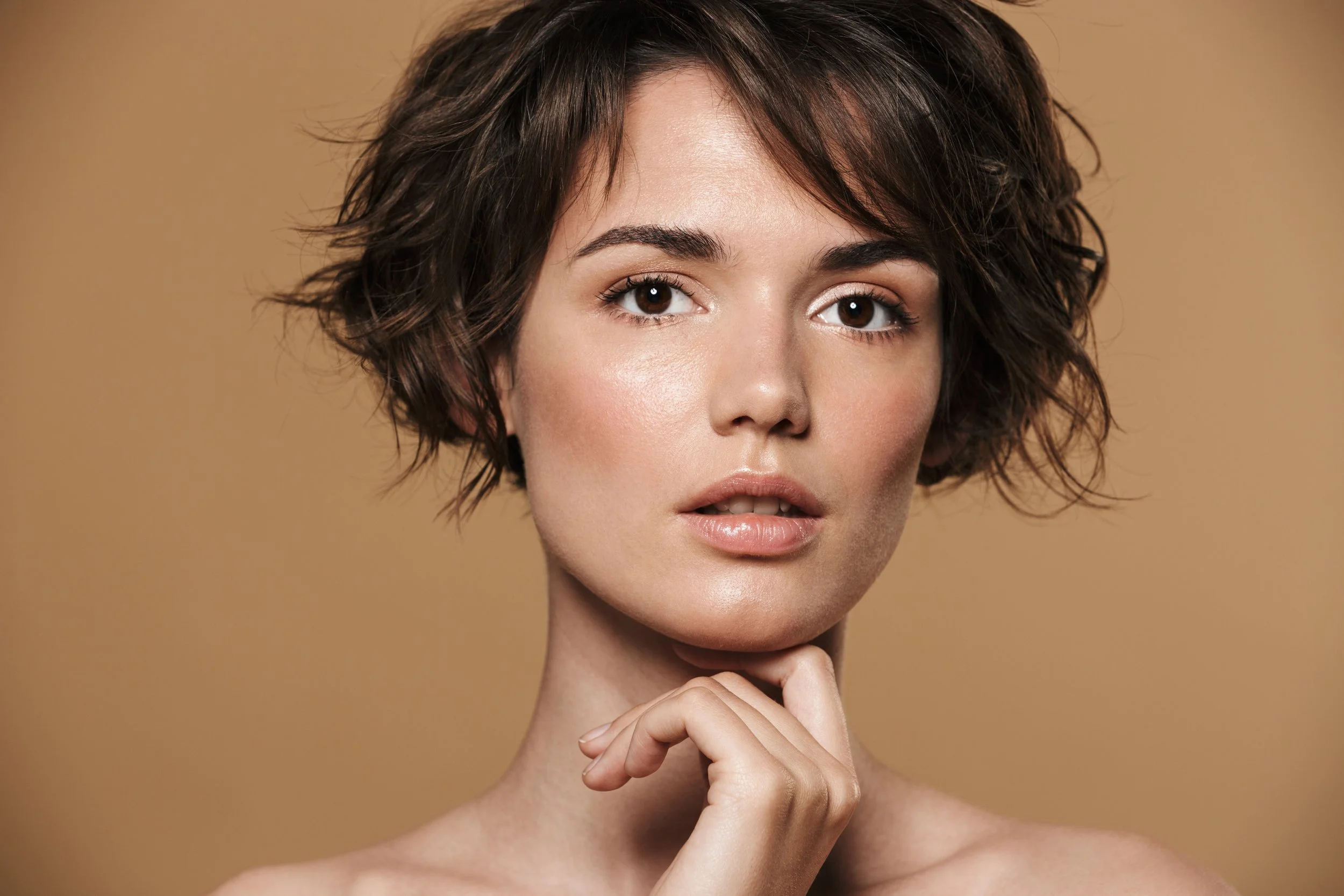 Close-up of a young woman with short, wavy brown hair, fair skin, and brown eyes, looking directly at the camera with a slightly open mouth, resting her chin on her hand against a beige background.