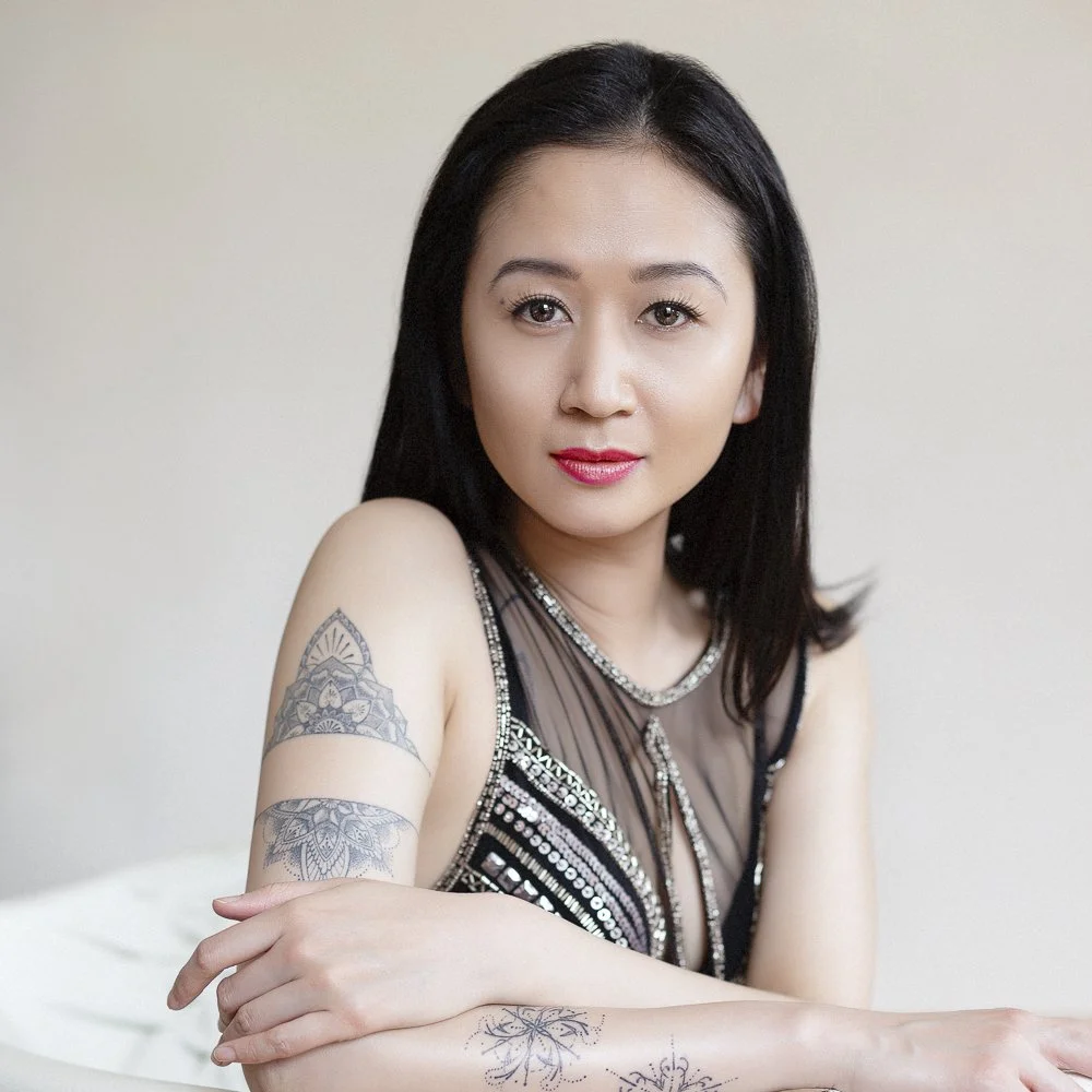 A woman with black hair and light skin is sitting with her arms crossed, showing tattoos on her arms, wearing a black dress with sequins and sheer fabric. She is looking at the camera with a slight smile.