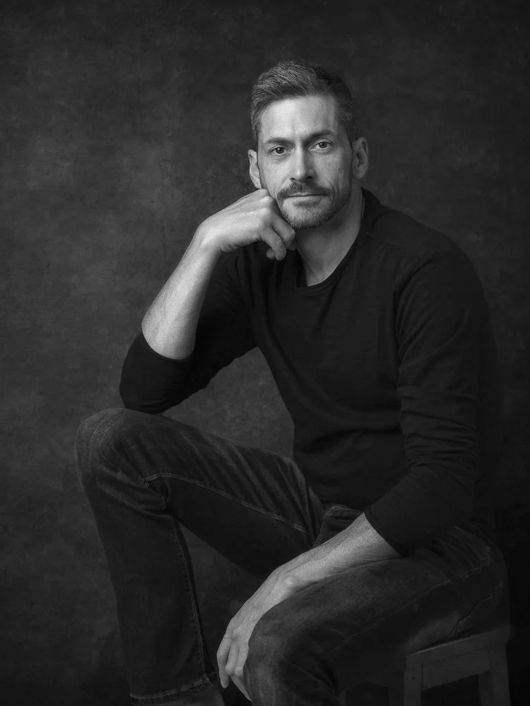 Black and white portrait of a man sitting on a stool, resting his chin on his hand, wearing a black long-sleeve shirt and jeans.
