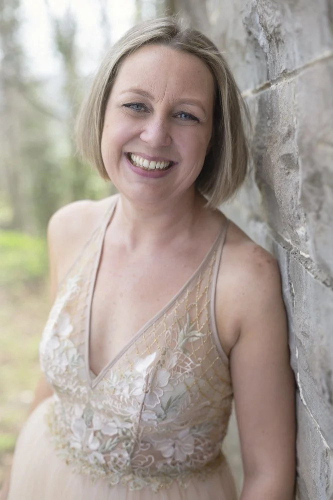 Smiling woman with short blonde hair and blue eyes leaning against a stone wall outdoors, wearing a sleeveless beige lace dress with floral embroidery.