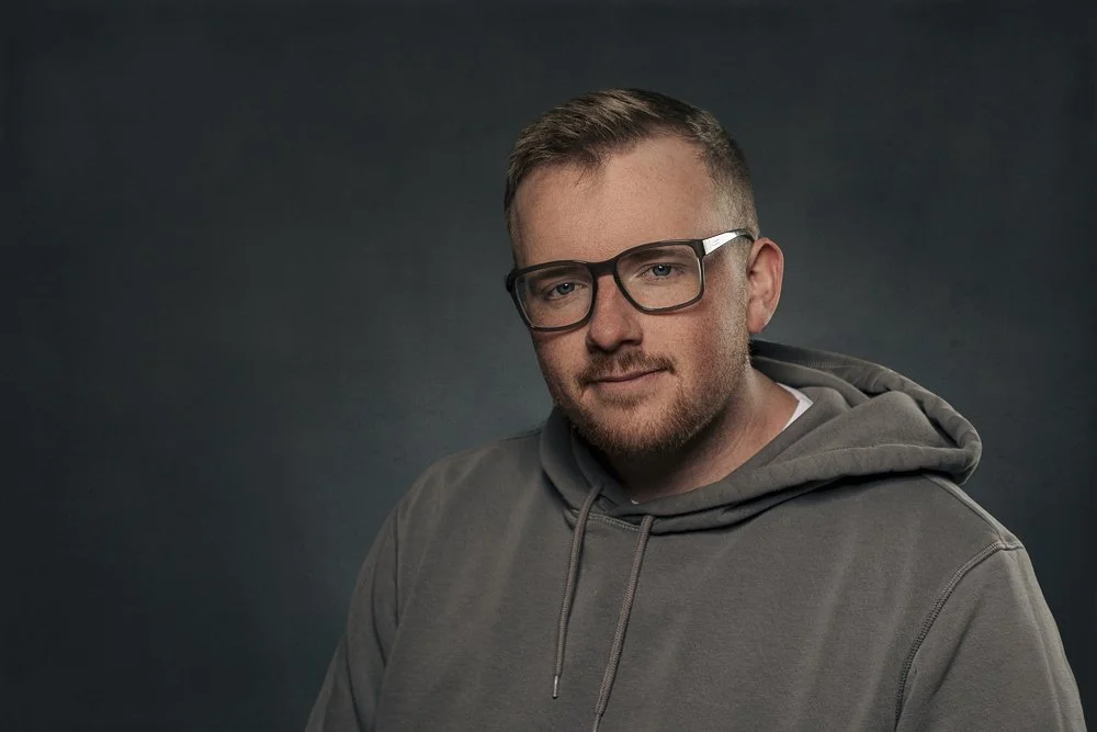 Portrait of a young man with short hair and beard, wearing glasses and a gray hoodie, against a dark background.