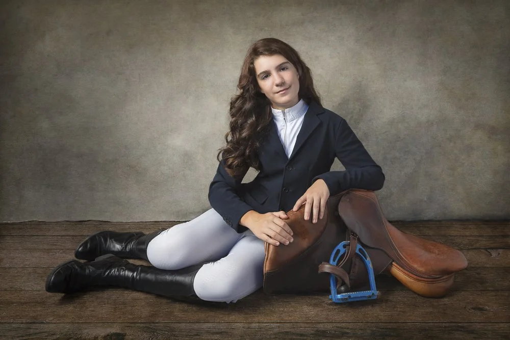 A young girl with long curly brown hair, wearing riding attire, sitting on wooden floor next to a horse saddle and stirrup.