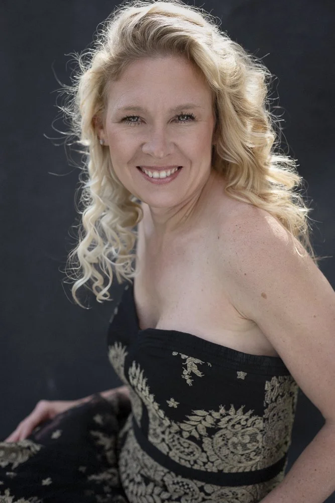 A smiling woman with blonde, curly hair, wearing a strapless black dress with gold floral patterns, against a dark background.