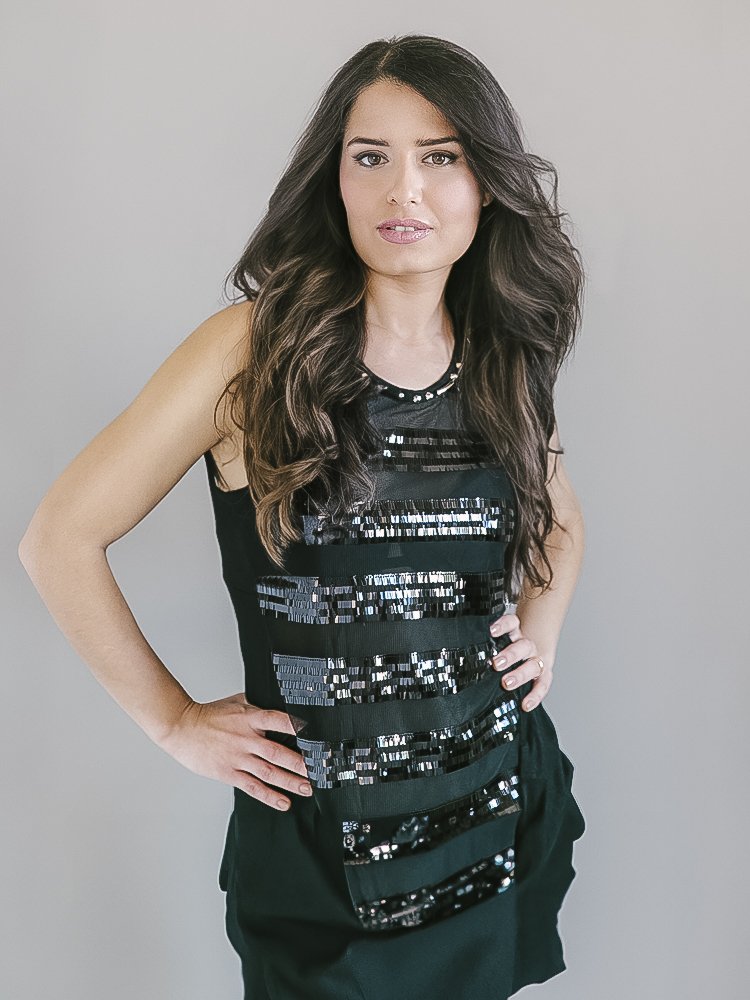 A woman with long dark hair in loose waves, wearing a sleeveless black top with horizontal shiny black sequin stripes, and black pants, standing against a plain gray background with her hand on her hip.
