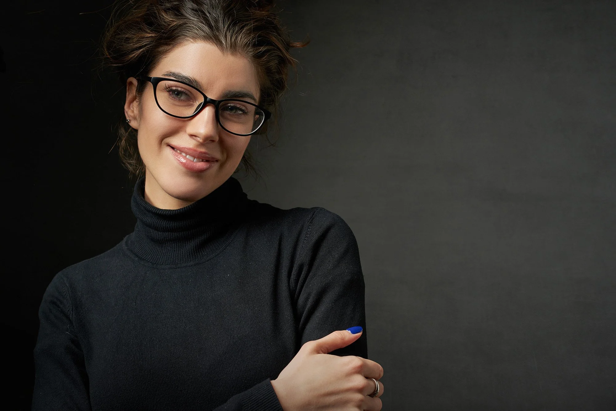 Young woman with short brown hair, wearing glasses and a black turtleneck, smiling and pointing to her left.