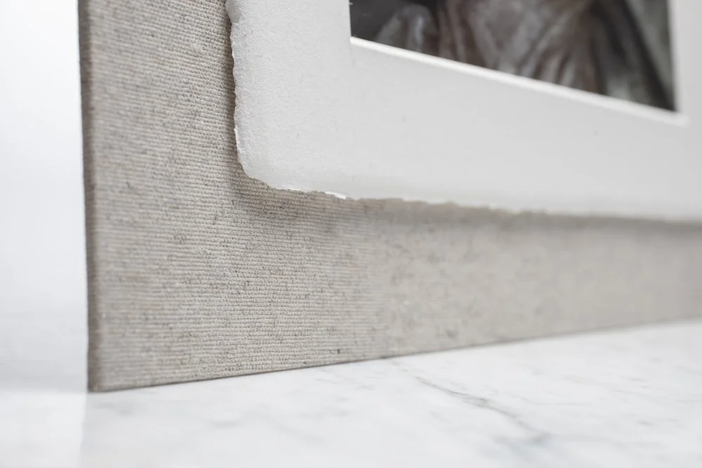 Close-up of a textured, beige fabric-covered picture frame with a glass window, sitting on a white marble surface.