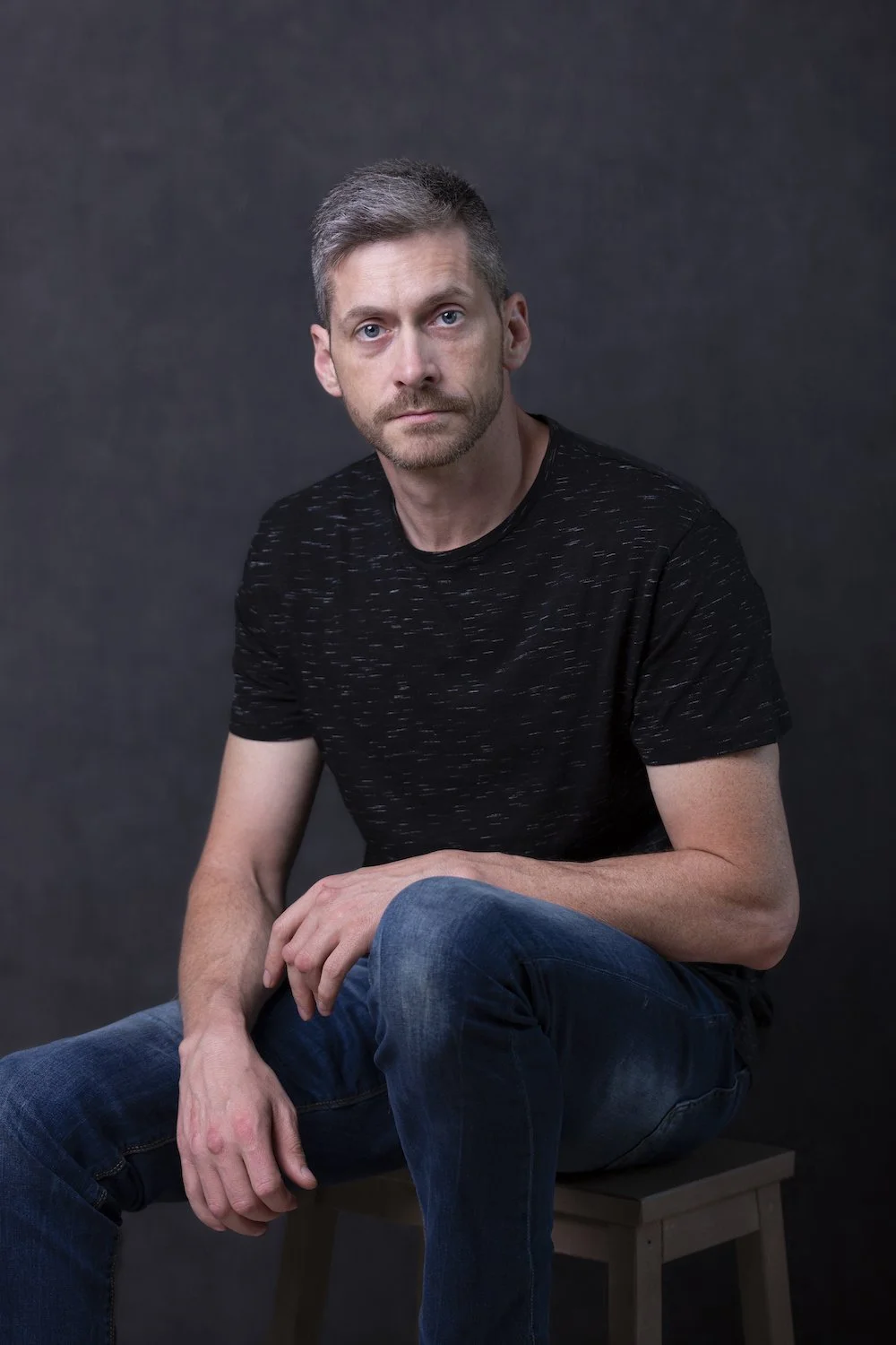 A man with gray hair and a beard wearing a black shirt with white streaks, sitting on a wooden stool against a dark background.