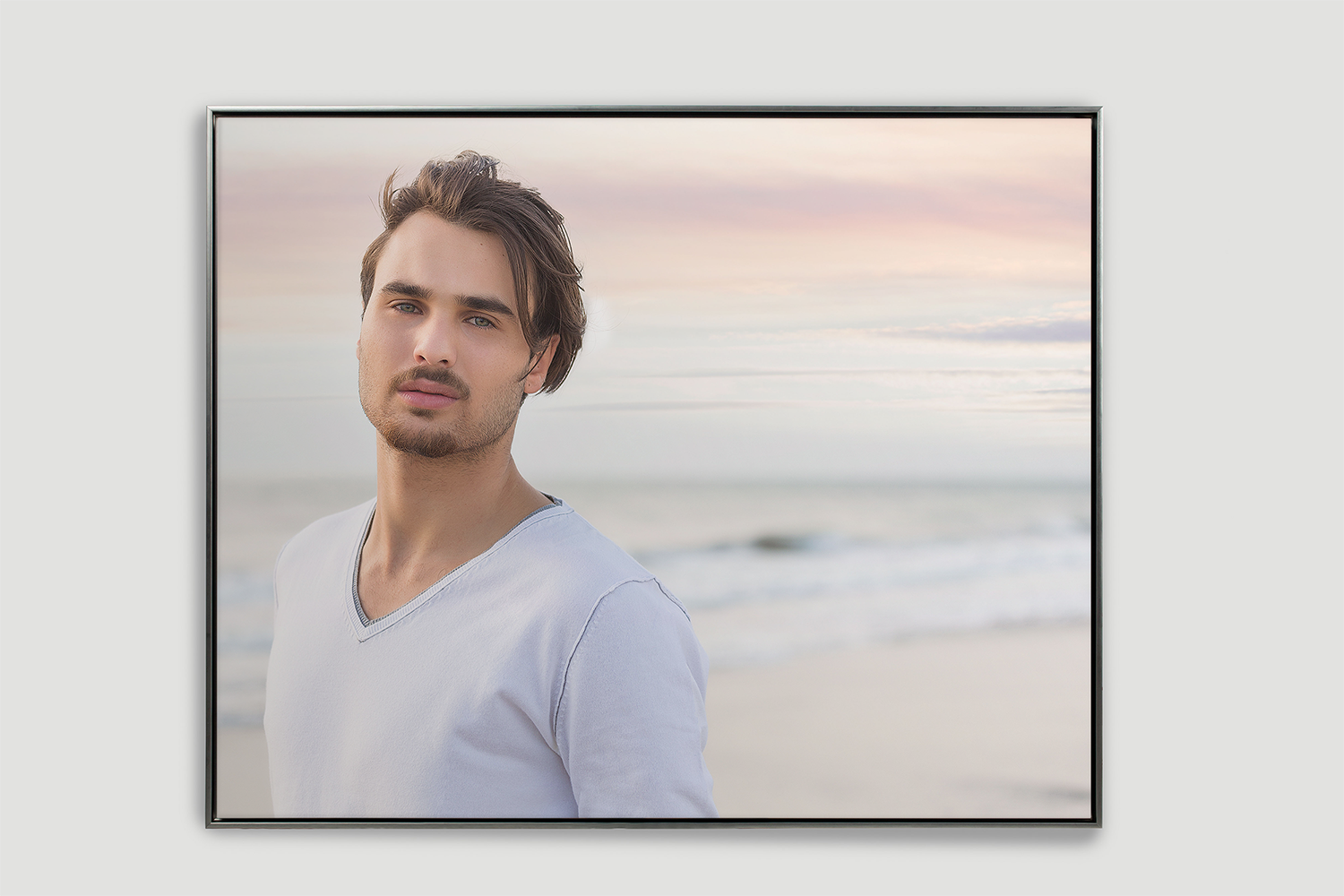A young man with dark hair, light beard, and light skin standing on a beach during sunset, wearing a white V-neck shirt, with ocean waves in the background.