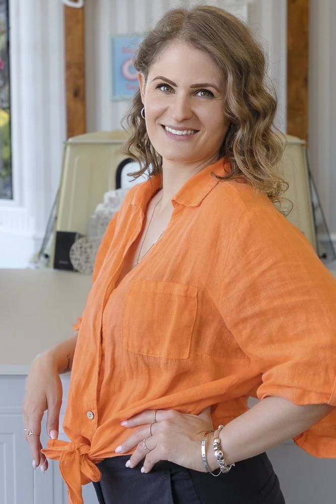 A woman with wavy, light brown hair, wearing an orange button-up shirt tied at the waist, standing indoors with a smile, in front of a white door and window, holding her waist with one hand.