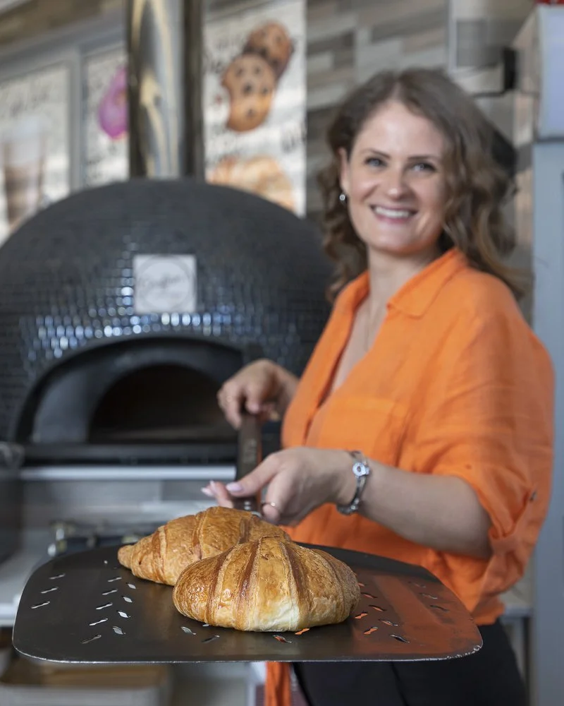A woman wearing an orange shirt holding a tray with two croissants in front of a pizza oven.