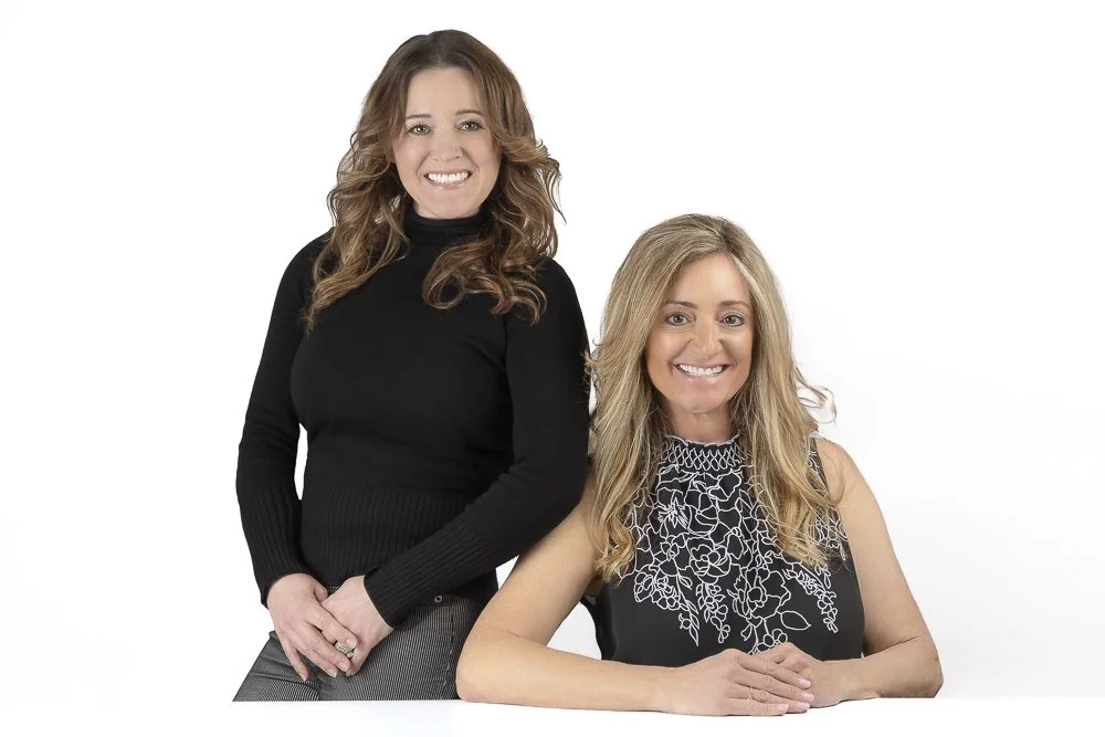 Two smiling women, one standing and one sitting, against a white background.