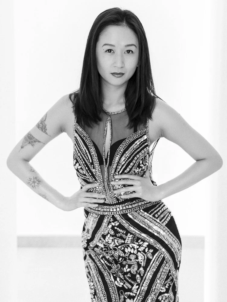 A woman with long dark hair, wearing a sleeveless, intricately patterned dress, standing with her hands on her hips.