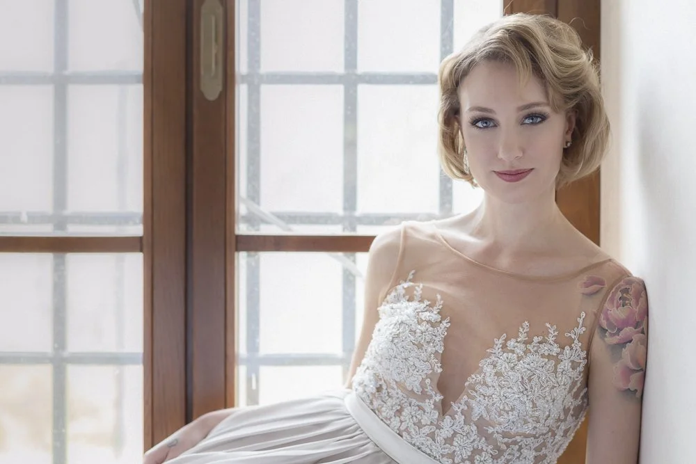 A woman with blonde hair in a lace dress sitting by a window with a wooden frame.