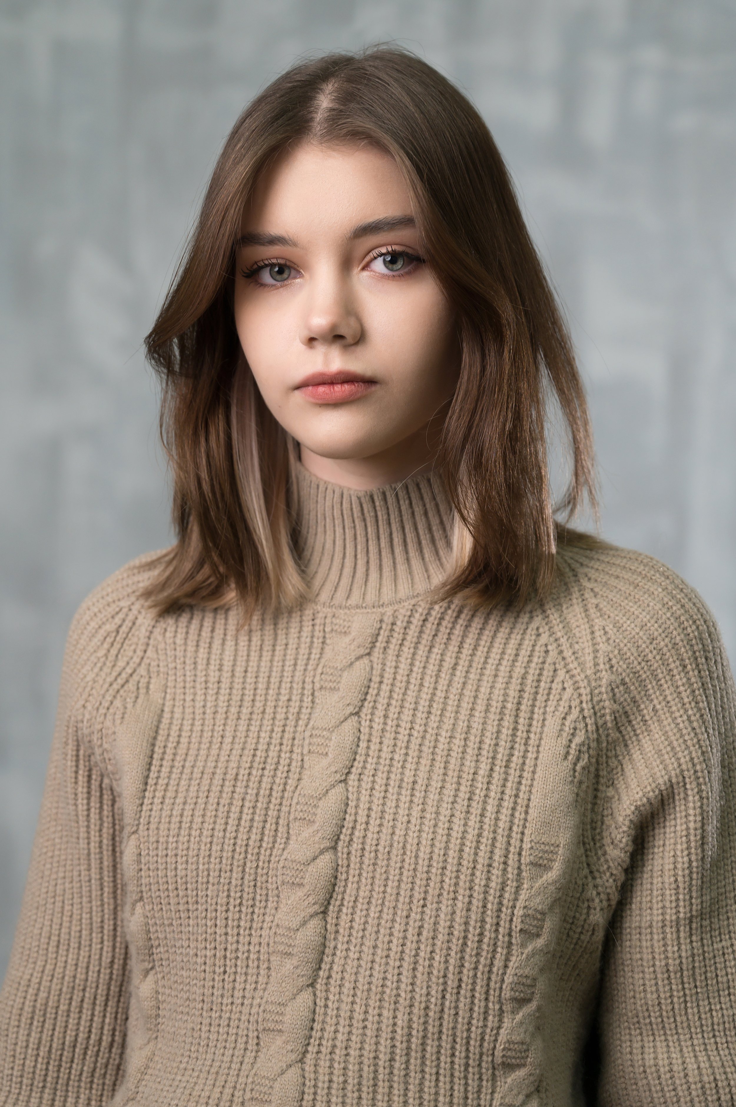 A young woman with shoulder-length brown hair and blue eyes, wearing a beige knit sweater, looking directly at the camera with a neutral expression against a gray textured background.