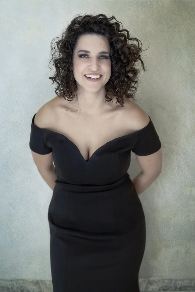A woman with curly dark hair smiling, wearing an off-the-shoulder black dress, standing against a light-colored wall.