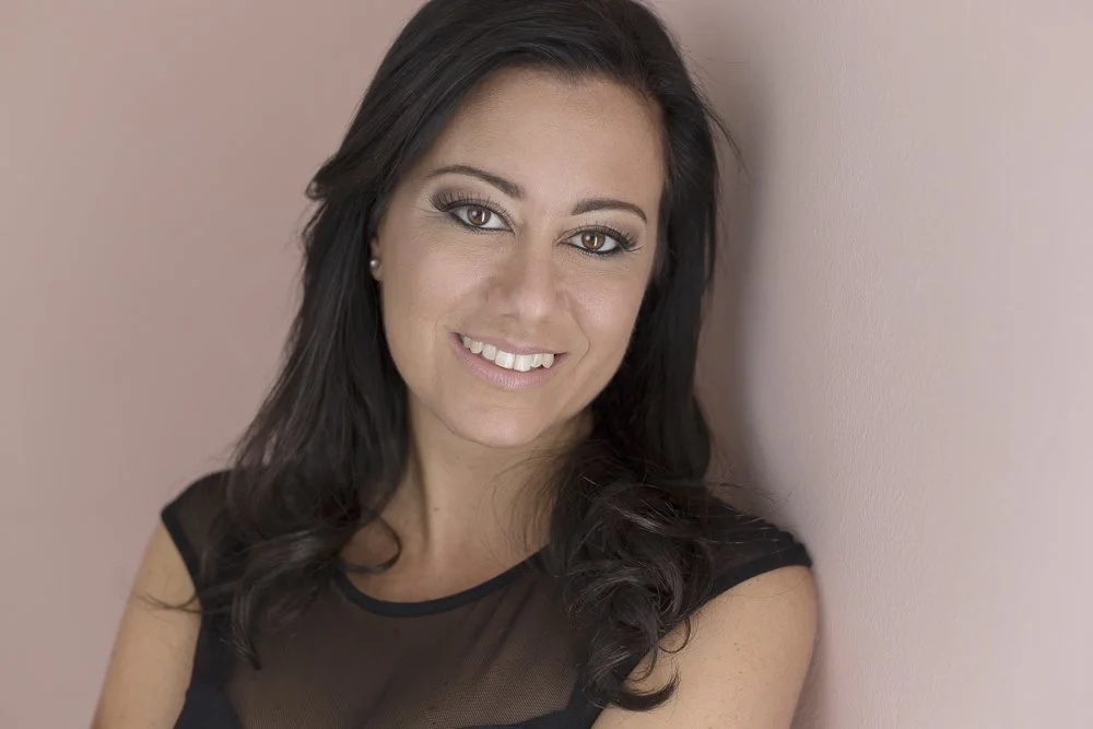 A woman with long dark hair smiling, leaning against a pink wall, wearing a black top.