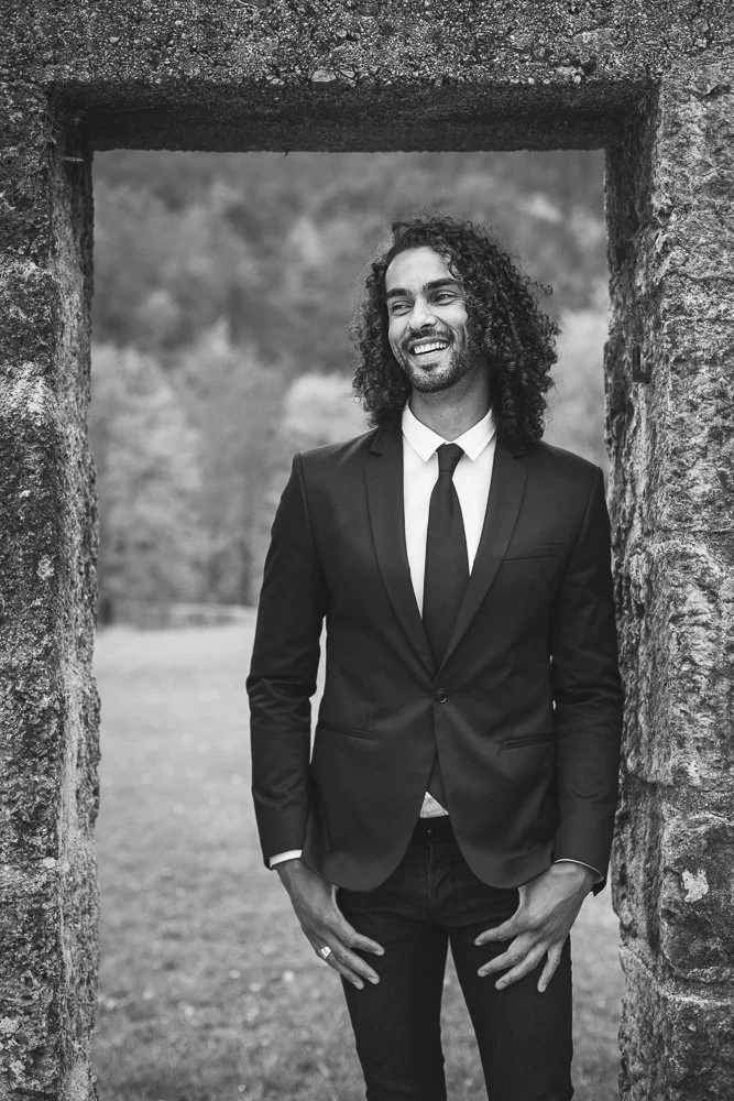 A man with curly hair and a beard, dressed in a black suit, white shirt, and black tie, smiling and standing outdoors within a stone doorway.