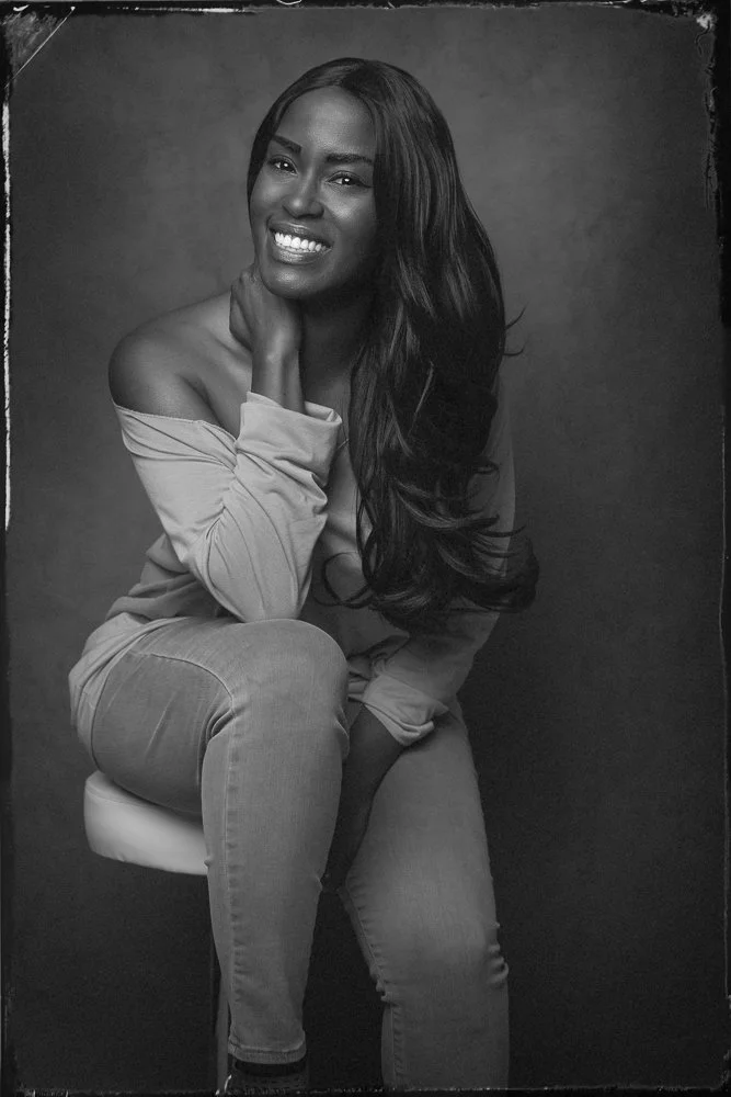 A woman with long, wavy hair sitting on a stool, smiling. She wears an off-the-shoulder top and jeans, with her hand resting on her neck. The photo is in black and white.