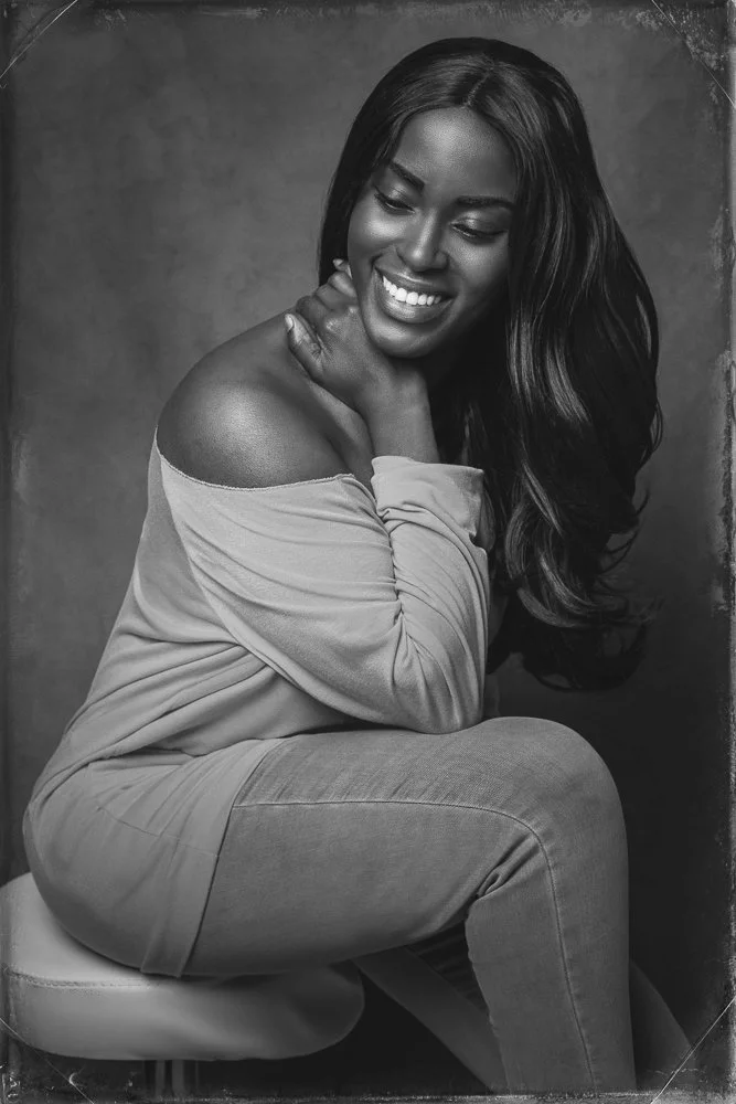 Black and white photo of a smiling woman with long hair, sitting on a chair, wearing a loose off-shoulder top and jeans, looking down.