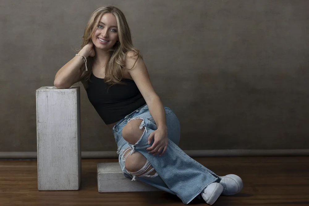 A young woman with long blonde hair wearing a black sleeveless top, ripped jeans, and white sneakers, smiling while sitting on the floor with her arm resting on a white rectangular block against a brown wall background.