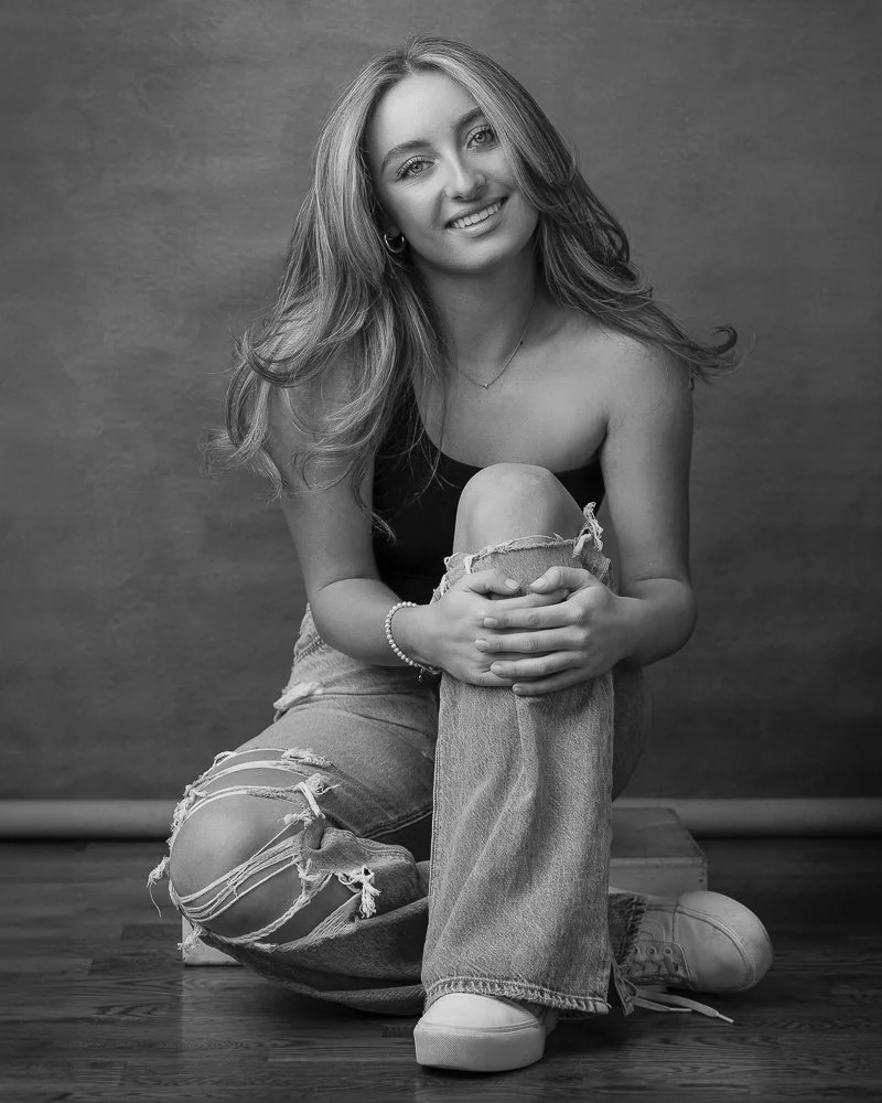 Black and white photo of a young woman smiling, sitting on the floor with one knee up, wearing ripped jeans, a sleeveless top, and sneakers, in front of a plain backdrop.