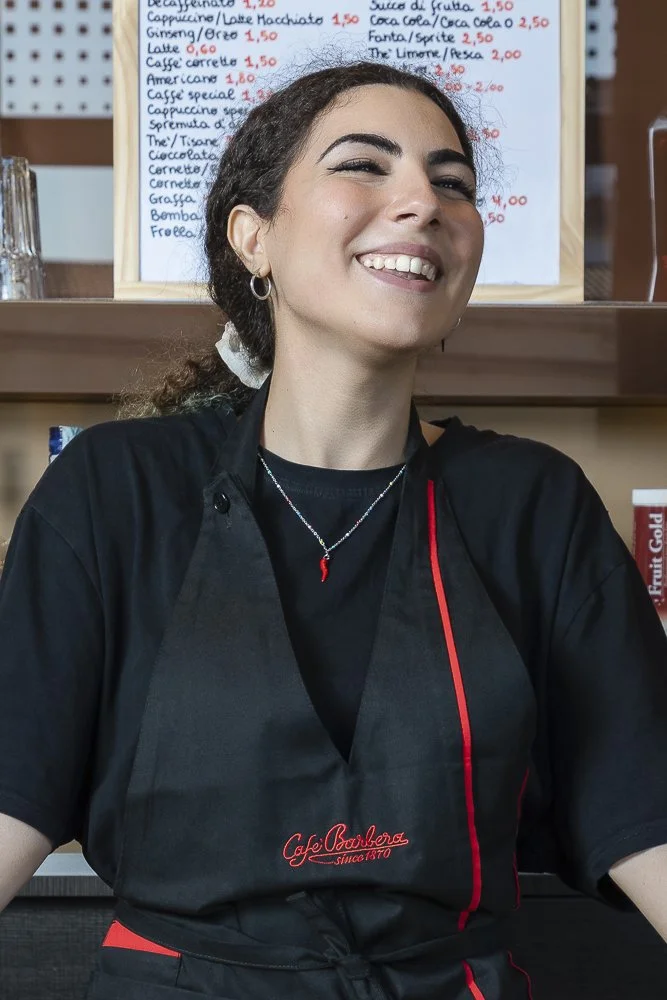 Smiling woman with dark hair tied back, wearing a black uniform with red accents and a small chili pepper necklace, standing in front of a menu board in a cafe.