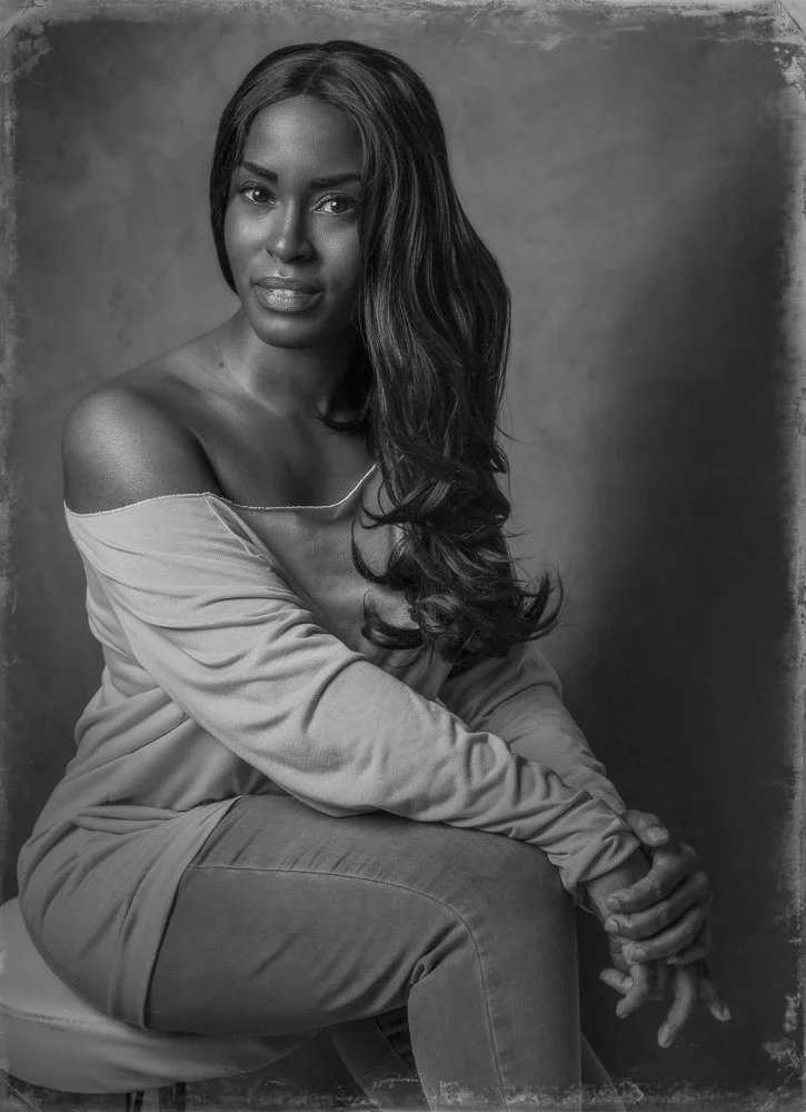 A woman with long, wavy hair sitting on a stool against a textured background, wearing an off-shoulder top and jeans, looking directly at the camera in a black-and-white portrait.