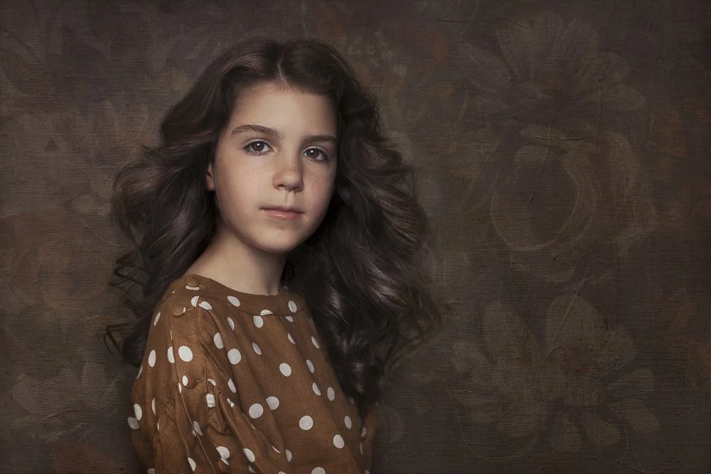 A young girl with long, curly brown hair wearing a brown polka-dot dress poses against a dark, textured background.