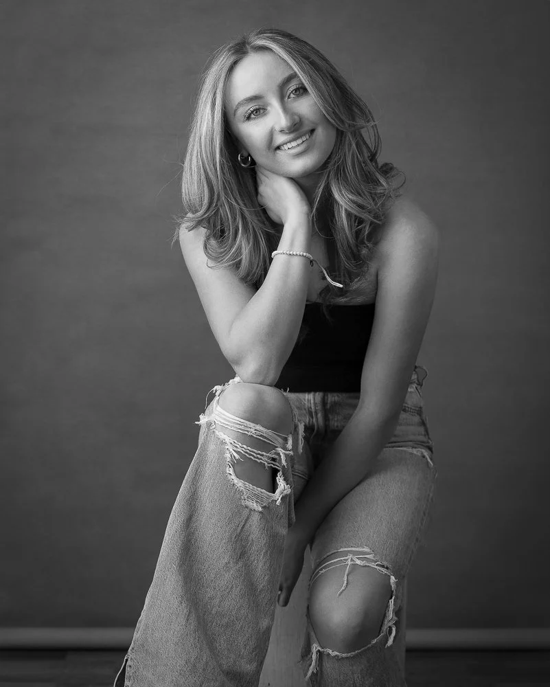 Black and white portrait of a young woman with wavy hair, smiling, sitting with one knee up. She is wearing a sleeveless top and ripped jeans, and resting her chin on her hand.