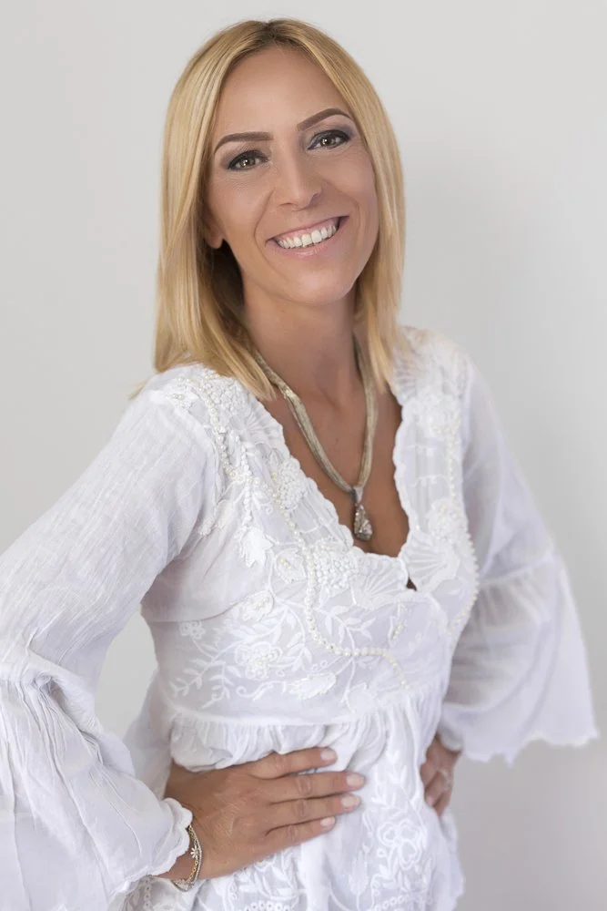 A smiling woman with shoulder-length blonde hair wearing a white embroidered blouse and a necklace, posing with her hand on her hip against a plain background.
