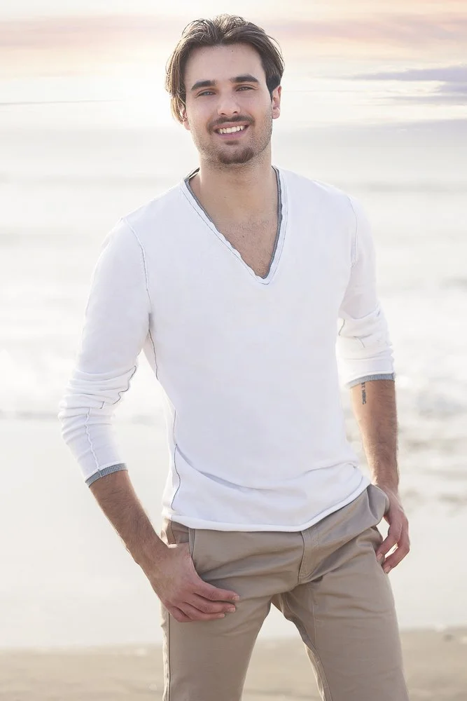 A young man with dark hair and a beard standing on a beach at sunset, smiling, wearing a white long-sleeve shirt and khaki pants.
