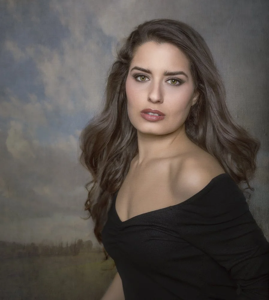 A young woman with long, wavy brown hair and green eyes wearing a black off-the-shoulder top, posing in front of a cloudy sky background.