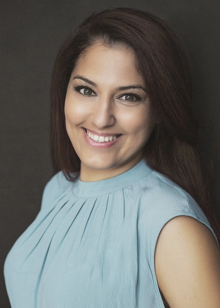 Close-up of a woman with brown hair, wearing a light blue sleeveless top, smiling against a dark background.