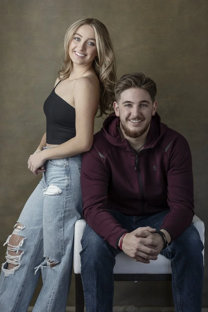A smiling young woman with long blonde hair wearing a black sleeveless top and ripped jeans, standing next to a young man with short brown hair and a beard, who is seated and wearing a maroon hoodie and jeans. They are against a plain background.