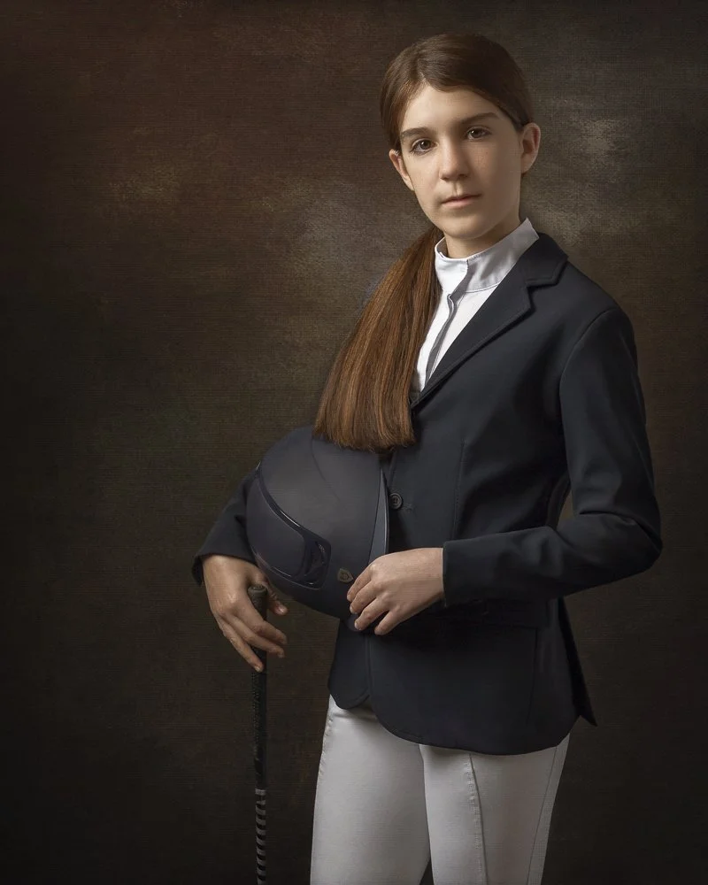 Young woman in a black blazer and white riding pants holding a black riding helmet and a riding crop against a dark background.