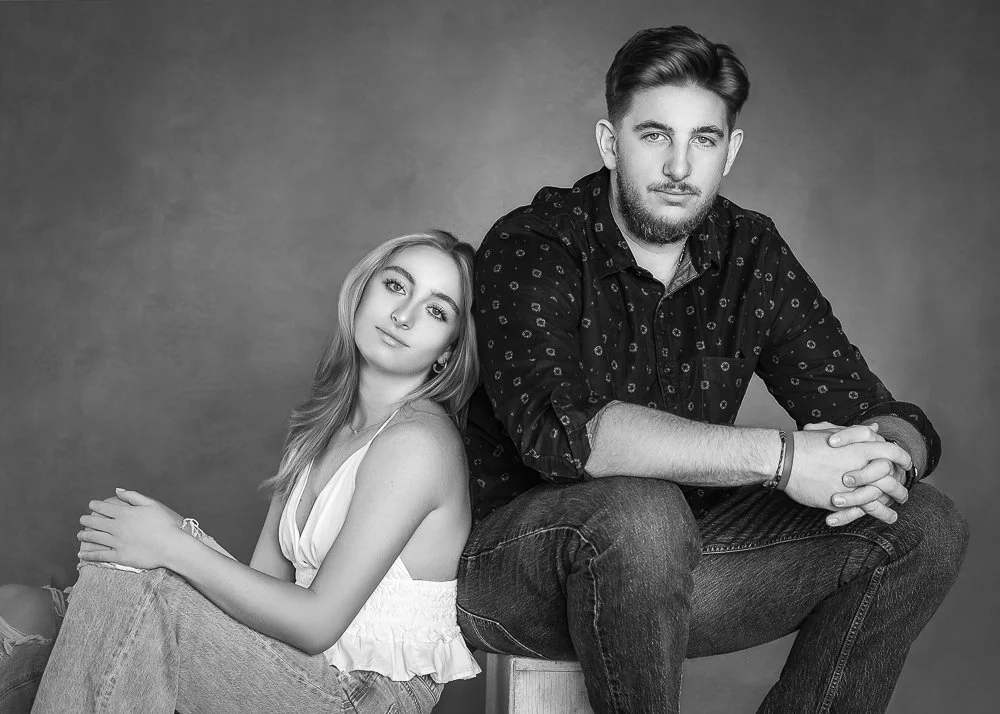 A black and white photo of a young woman and man posing together. The woman is leaning on the man's shoulder, and they are both looking at the camera. The woman has long hair and is wearing a sleeveless top, while the man has styled hair, a beard, an
