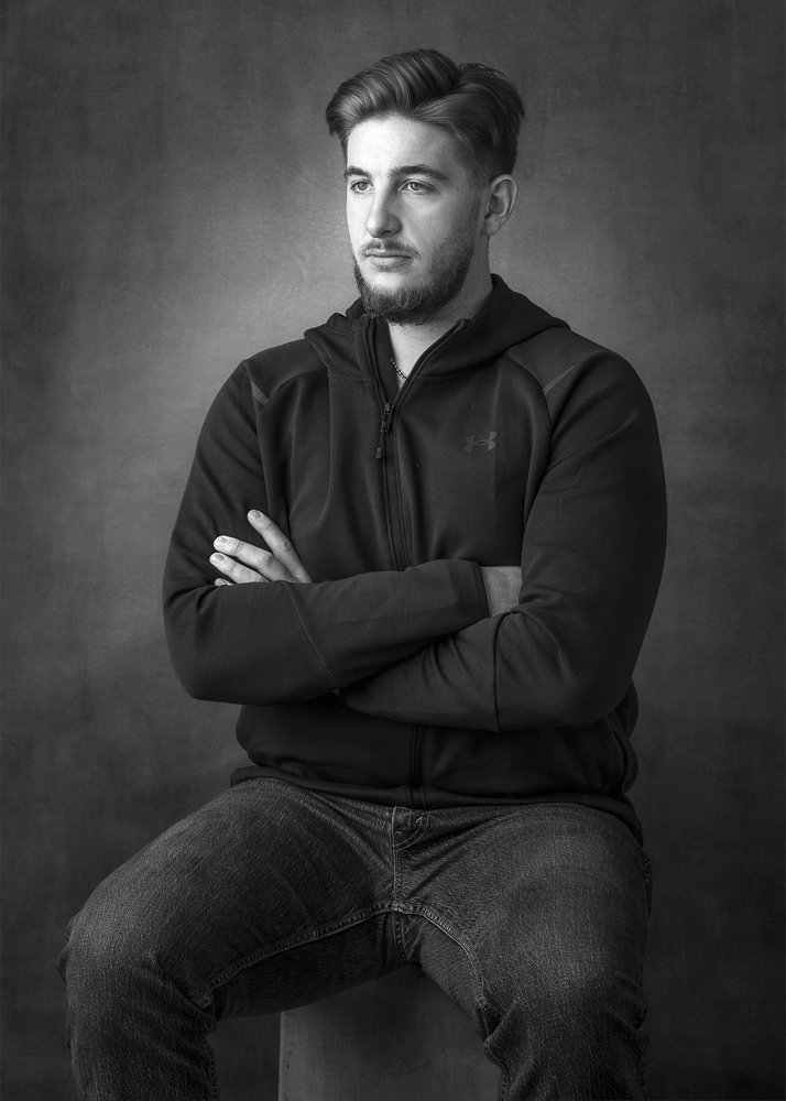 Black and white portrait of a young man with styled hair, a beard, wearing a hoodie and jeans, sitting and looking to the side with arms crossed.