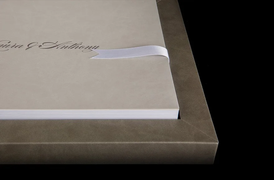 A beige wedding invitation with elegant black cursive text, partially visible, inside a brown box with a white ribbon.