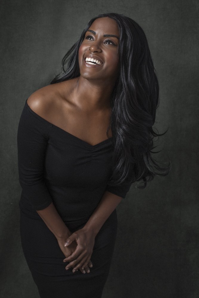 Portrait of a smiling Black woman with long wavy black hair, wearing a black off-shoulder dress, standing against a dark textured background.
