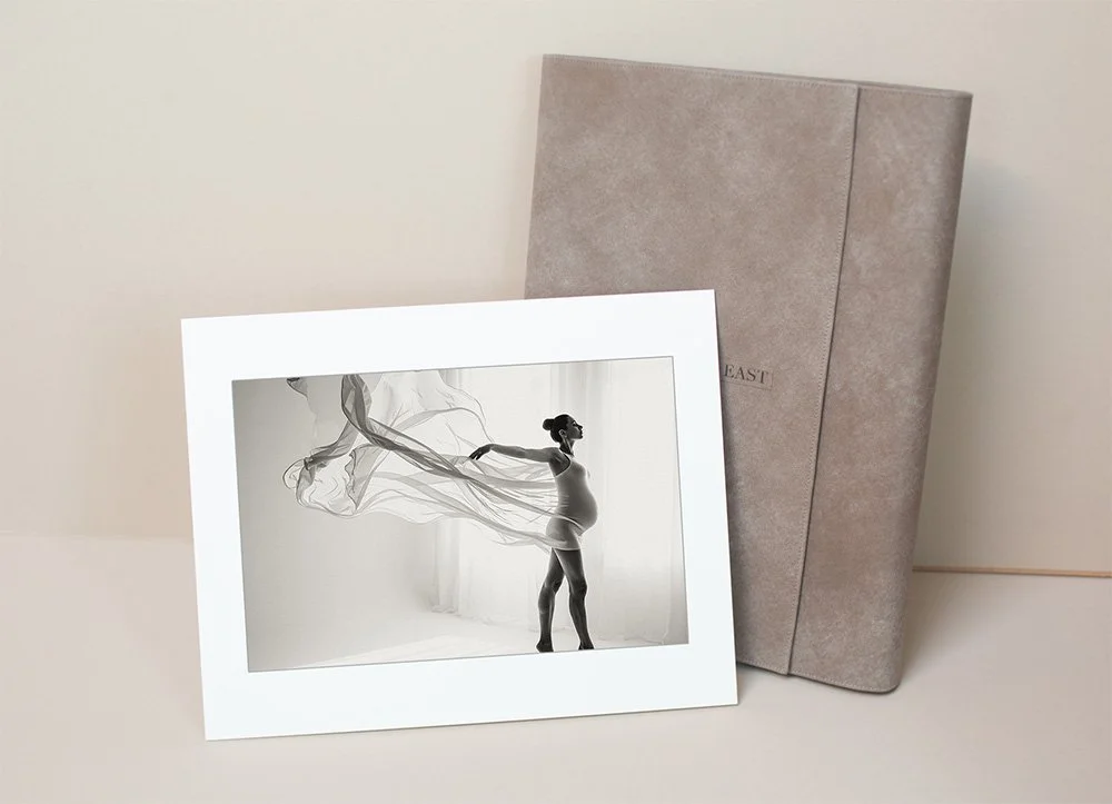 A framed black-and-white photograph of a pregnant woman standing by a window, with flowing sheer fabric around her, is leaning against a beige textured box or cushion.