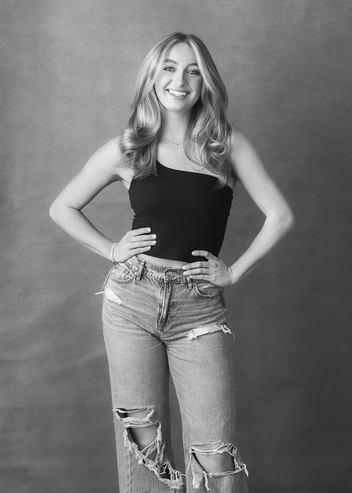 A young woman with long wavy hair smiling and posing with her hands on her hips, wearing a sleeveless black top and ripped jeans, against a plain background.