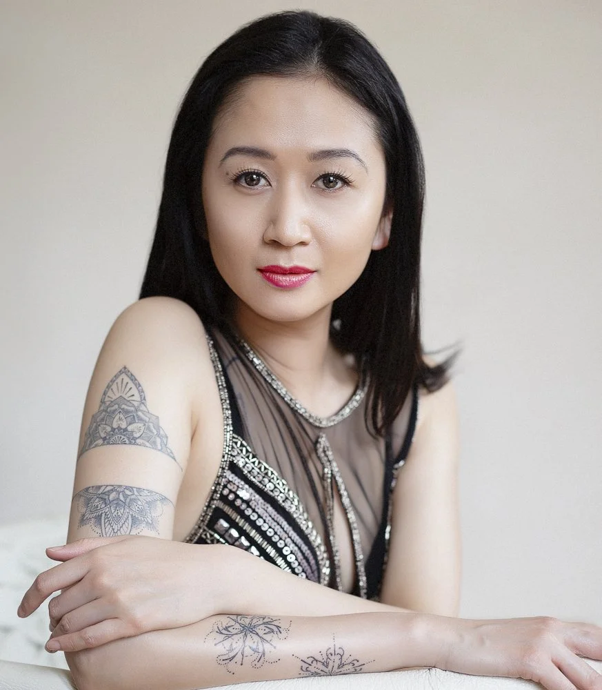 A woman with straight black hair and tattoos on her arm, wearing a sleeveless black dress with silver embellishments, sitting and looking at the camera.