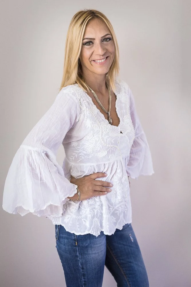 A smiling woman with long blonde hair wearing a white embroidered blouse with ruffled sleeves and blue jeans.